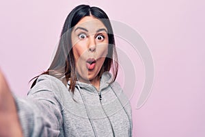 Young beautiful brunette sporty woman make selfie by the camera over pink background scared and amazed with open mouth for