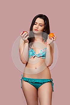 A young beautiful brunette woman in a blue swimsuit with fresh oranges and apples in her hands on a pink isolated background