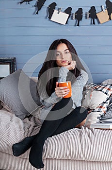 Young beautiful brunette woman with cup of coffee sitting home on the sofa by the window.