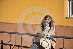 Young, beautiful, brunette woman with curly hair and beige coat and hat, leaning on a railing looking at camera thoughtful and