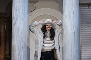 Young, beautiful, brunette woman with curly hair, beige sweater, coat and hat, leaning on marble columns, smiling and happy.