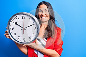 Young beautiful brunette woman doing countdown holding big clock over blue background looking positive and happy standing and