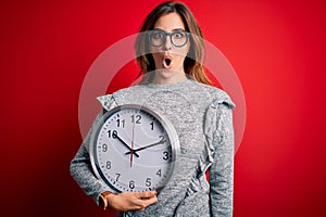 Young beautiful brunette woman doing countdown holding big clock over red background scared in shock with a surprise face, afraid