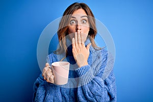 Young beautiful brunette woman drinking cup of coffe over isolated blue background cover mouth with hand shocked with shame for