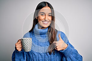 Young beautiful brunette woman drinking cup of coffee over isolated white background happy with big smile doing ok sign, thumb up