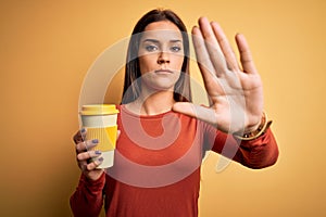 Young beautiful brunette woman drinking cup of takeaway coffe over yellow background with open hand doing stop sign with serious