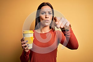 Young beautiful brunette woman drinking cup of takeaway coffe over yellow background pointing with finger to the camera and to
