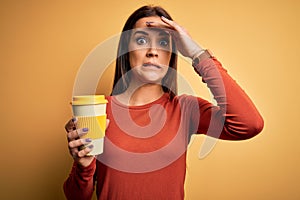 Young beautiful brunette woman drinking cup of takeaway coffe over yellow background stressed with hand on head, shocked with