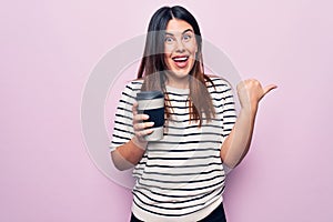 Young beautiful brunette woman drinking cup of takeaway coffee over isolated pink background pointing thumb up to the side smiling