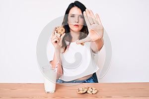 Young beautiful brunette woman drinking a glass of fresh milk with cookies with open hand doing stop sign with serious and