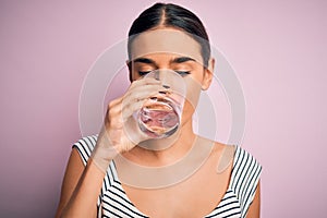 Young beautiful brunette woman drinking glass of healthy water to refreshment standing over isolated pink background