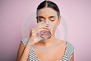 Young beautiful brunette woman drinking glass of healthy water to refreshment standing over isolated pink background