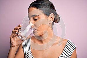 Young beautiful brunette woman drinking glass of healthy water to refreshment standing over isolated pink background