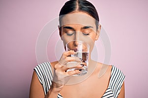 Young beautiful brunette woman drinking glass of healthy water to refreshment standing over isolated pink background