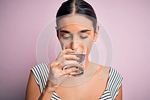 Young beautiful brunette woman drinking glass of healthy water to refreshment standing over isolated pink background