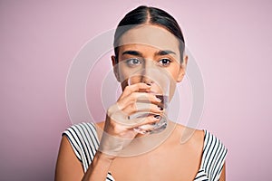 Young beautiful brunette woman drinking glass of healthy water to refreshment standing over isolated pink background