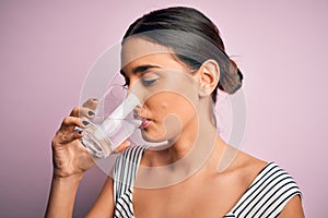 Young beautiful brunette woman drinking glass of healthy water to refreshment standing over isolated pink background