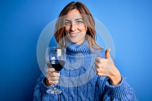 Young beautiful brunette woman drinking glass of red wine over  blue background happy with big smile doing ok sign, thumb