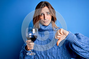 Young beautiful brunette woman drinking glass of red wine over isolated blue background with angry face, negative sign showing