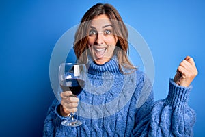 Young beautiful brunette woman drinking glass of red wine over isolated blue background screaming proud and celebrating victory