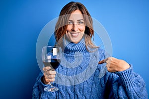 Young beautiful brunette woman drinking glass of red wine over isolated blue background with surprise face pointing finger to