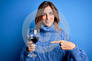 Young beautiful brunette woman drinking glass of red wine over isolated blue background very happy pointing with hand and finger