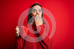 Young beautiful brunette woman drinking mug of coffe over isolated red background cover mouth with hand shocked with shame for