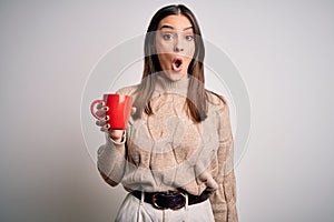 Young beautiful brunette woman drinking red cup of coffee over isolated white background scared in shock with a surprise face,