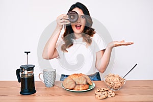 Young beautiful brunette woman eating breakfast holding cholate donut celebrating achievement with happy smile and winner