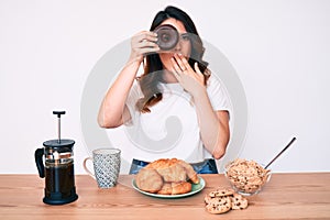 Young beautiful brunette woman eating breakfast holding cholate donut covering mouth with hand, shocked and afraid for mistake