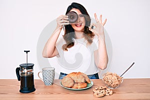 Young beautiful brunette woman eating breakfast holding cholate donut doing ok sign with fingers, smiling friendly gesturing