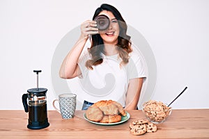 Young beautiful brunette woman eating breakfast holding cholate donut looking positive and happy standing and smiling with a