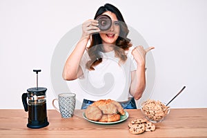 Young beautiful brunette woman eating breakfast holding cholate donut pointing thumb up to the side smiling happy with open mouth