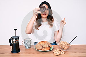 Young beautiful brunette woman eating breakfast holding cholate donut smiling happy pointing with hand and finger to the side
