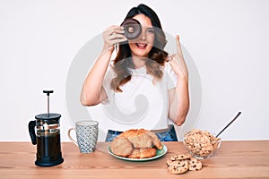 Young beautiful brunette woman eating breakfast holding cholate donut smiling with an idea or question pointing finger with happy