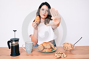 Young beautiful brunette woman eating breakfast holding croissant with open hand doing stop sign with serious and confident