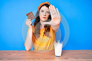 Young beautiful brunette woman eating a chocolate bar and drinking glass of milk with open hand doing stop sign with serious and