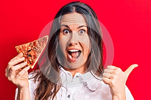 Young beautiful brunette woman eating slice of Italian pizza over isolated red background pointing thumb up to the side smiling