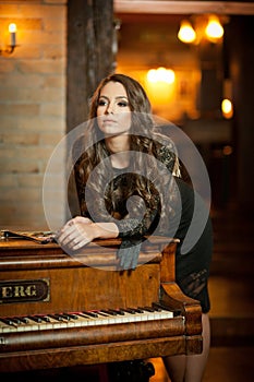 Young beautiful brunette woman in elegant black dress standing near a vintage piano. Sensual romantic lady with long dark hair