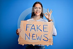 Young beautiful brunette woman holding banner with fake news message doing ok sign with fingers, excellent symbol