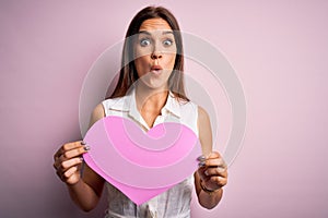 Young beautiful brunette woman holding big pink heart paper celebrating valentine day scared in shock with a surprise face, afraid