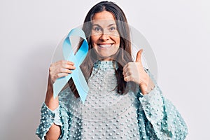 Young beautiful brunette woman holding blue ribbon symbol over isolated white background smiling happy and positive, thumb up