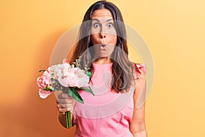 Young beautiful brunette woman holding bouquet of pink flowers over yellow background scared and amazed with open mouth for
