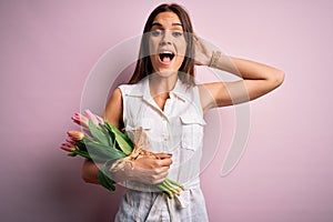 Young beautiful brunette woman holding bouquet of tulips flowers over pink background Crazy and scared with hands on head, afraid