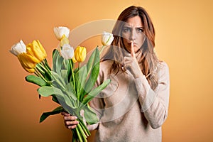 Young beautiful brunette woman holding bouquet of yellow tulips over  background asking to be quiet with finger on lips