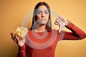 Young beautiful brunette woman holding bowl with chips potatoes over yellow background with angry face, negative sign showing