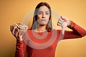 Young beautiful brunette woman holding bowl with cornflakes cereals over yellow background with angry face, negative sign showing