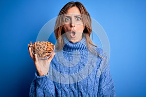 Young beautiful brunette woman holding bowl german baked pretzels over blue background scared in shock with a surprise face,