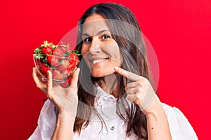 Young beautiful brunette woman holding bowl of heallthy strawberries over red background smiling happy pointing with hand and