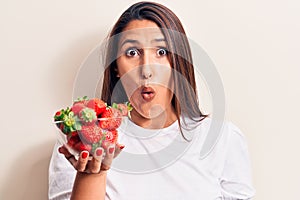Young beautiful brunette woman holding bowl with strawberries scared and amazed with open mouth for surprise, disbelief face